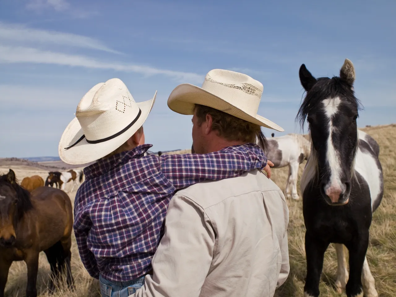 Son has his arm around father's neck, both in cowboy hats looking out at horses